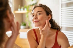 Beauty girl looking at mirror while touching her face and checking pimple, wrinkles and bags under the eyes, during morning beauty routine. Happy smiling beautiful young woman applying moisturiser.